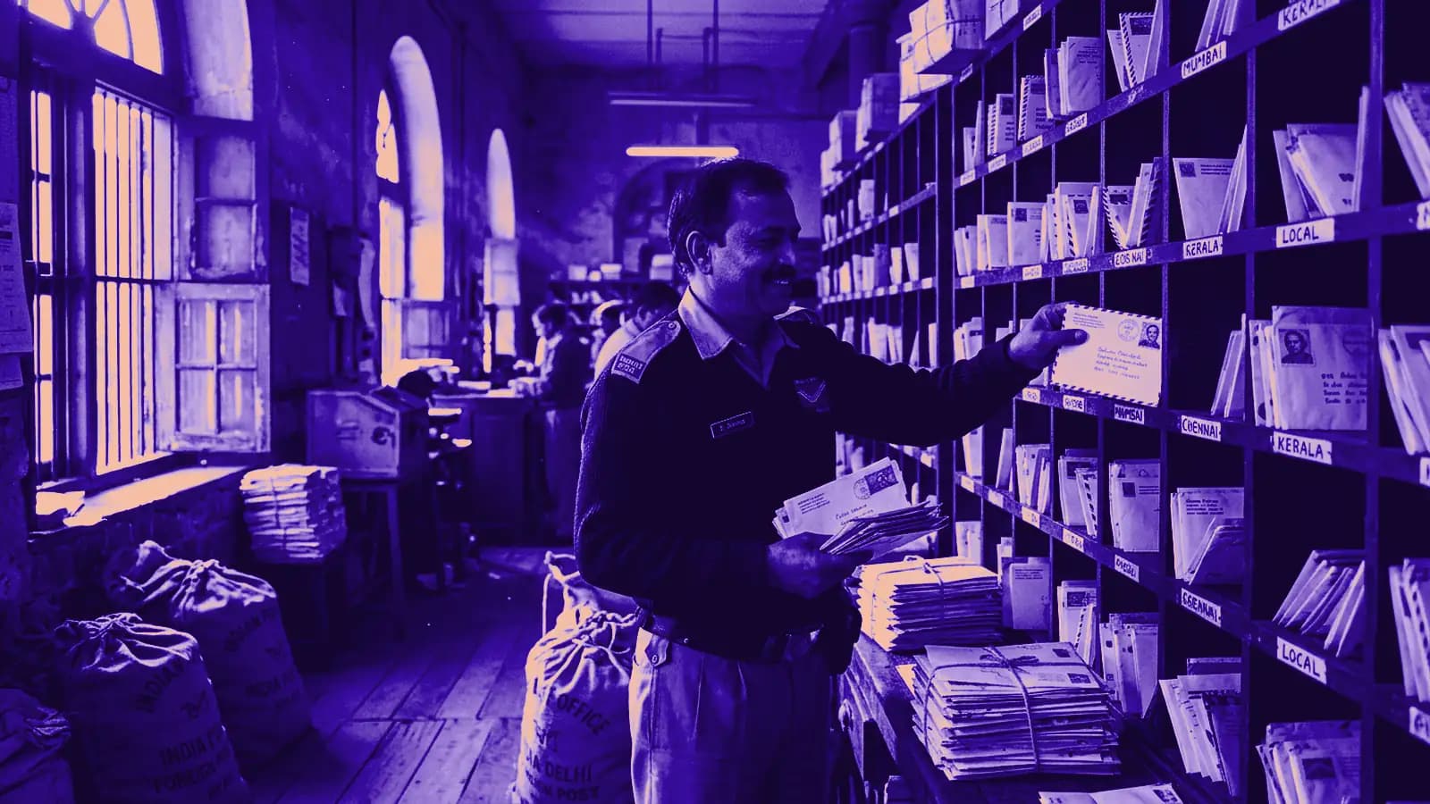 Postman sorting stamped envelopes into wooden pigeonhole racks in head post office mail sorting room with morning window dust motes